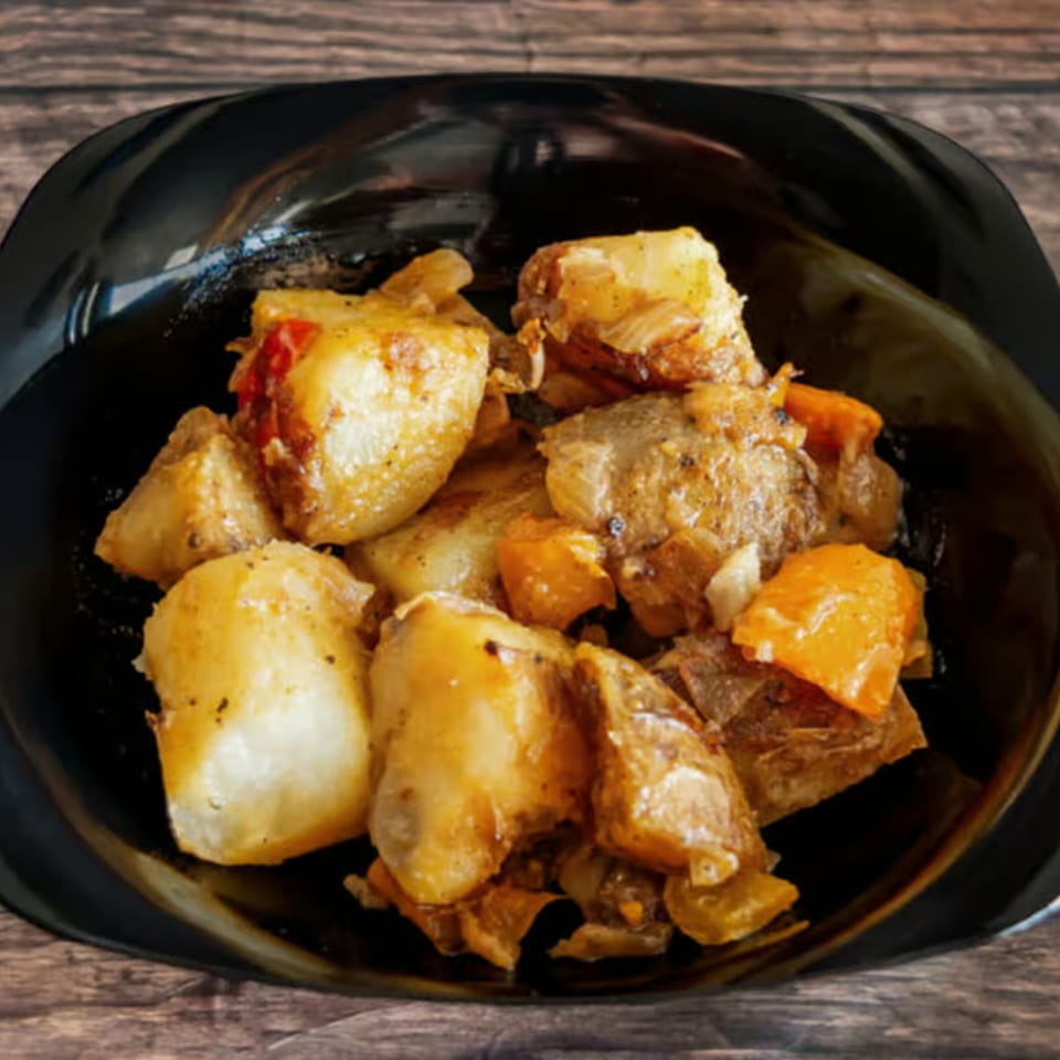 A black bowl filled with roasted potatoes, onions, and orange bell peppers on a wooden surface.