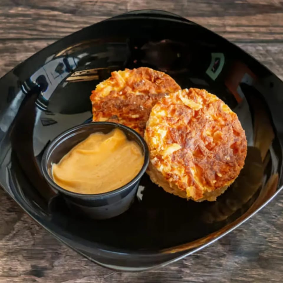 Two golden-brown hash browns and a small cup of dipping sauce on a black plate.